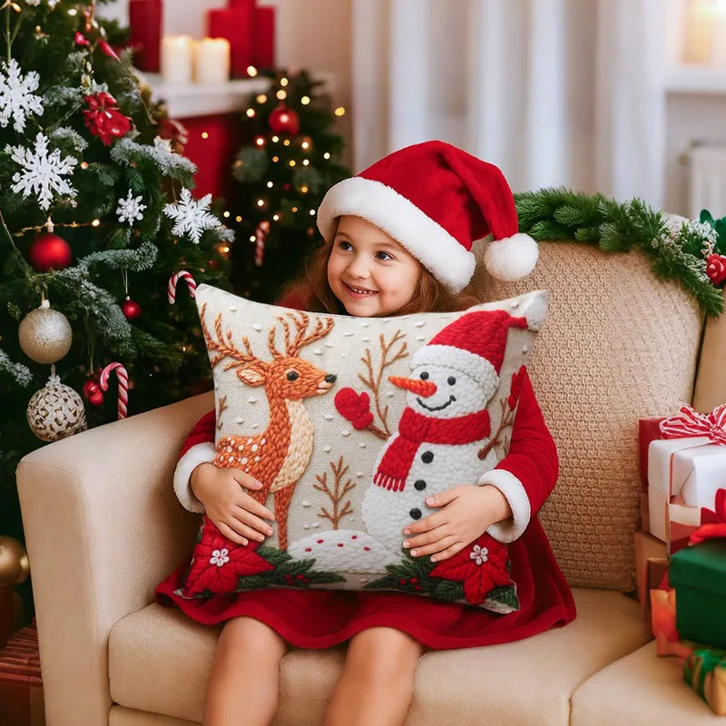 Child in a red Santa hat holding a Christmas-themed pillow with a reindeer and snowman design, surrounded by festive decorations.
