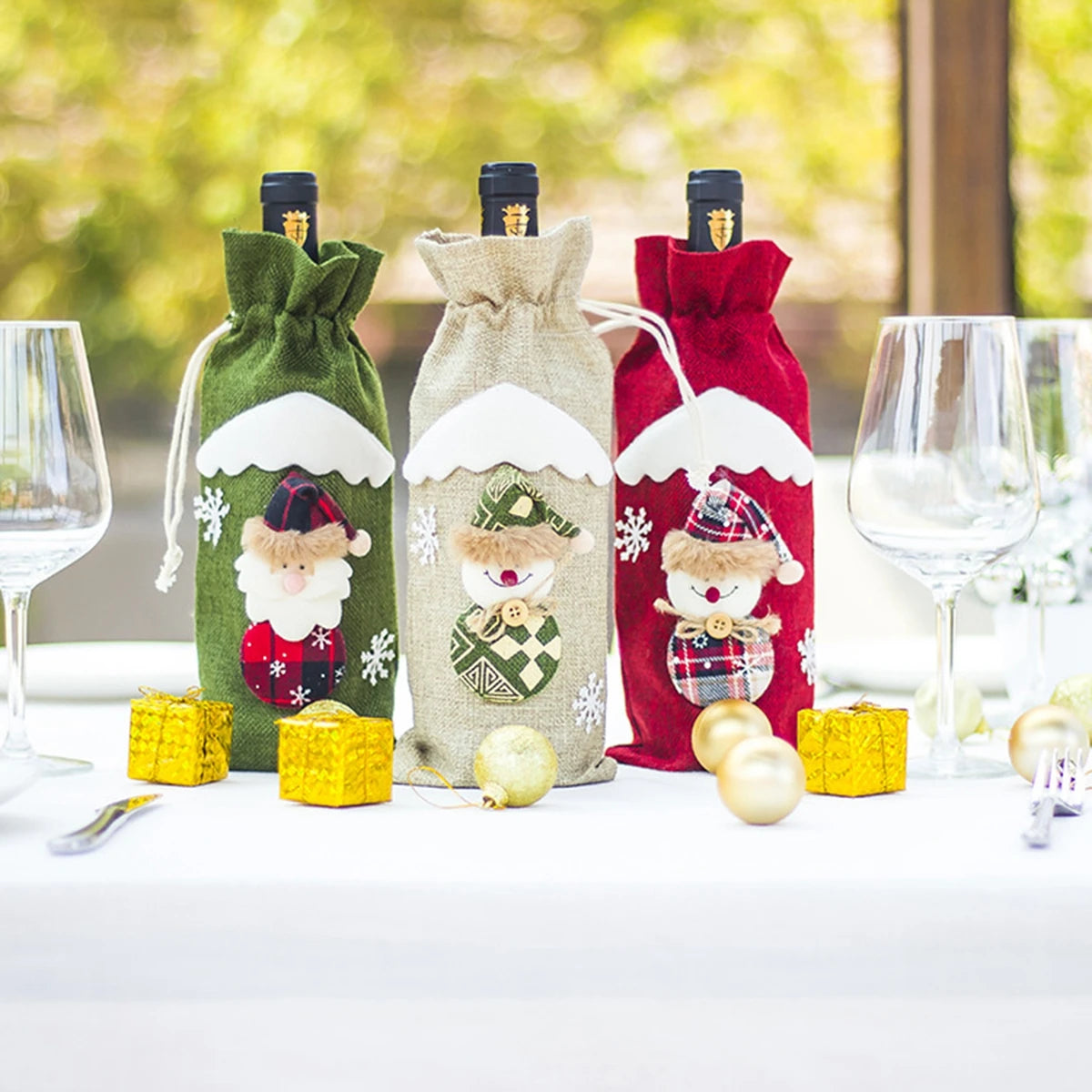 Three Christmas-themed wine bottle covers on a table with glasses and ornaments.