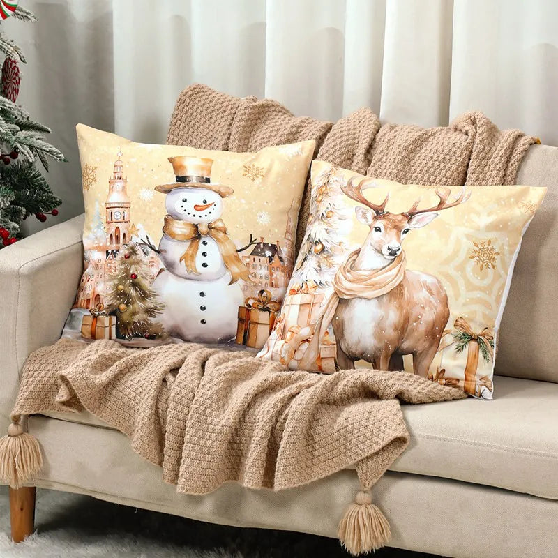 Decorative pillows with a snowman and deer design on a beige sofa with a Christmas tree in the background.