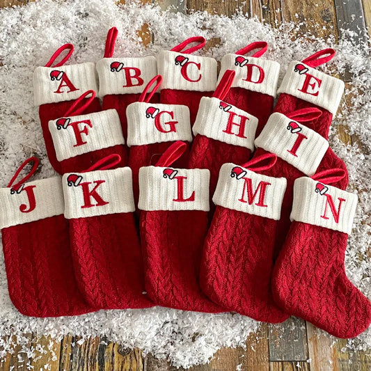 Red and white Christmas stockings with letters A to N on a wooden surface with snow-like texture.